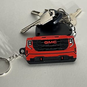 May include: A red and black GMC truck-shaped keychain with a silver keyring. The keychain is a miniature replica of a truck's front grille, with the GMC logo prominently displayed. Several keys and a key fob are also present.