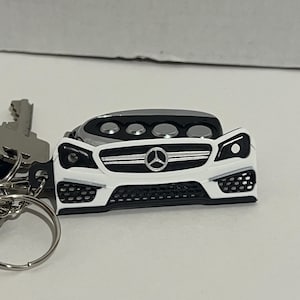 Op de afbeelding: Een wit en zwart auto-vormige sleutelhanger met een zilveren sleutel en sleutelring. De sleutelhanger heeft een auto-ontwerp met een zwarte grille, koplampen en het Mercedes-Benz-logo. Het dak van de auto is grijs.
