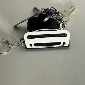 May include: A white car-shaped keychain with a black border and a silver keyring. The keychain is next to a set of car keys on a light gray surface. The car keychain features a detailed front grill and headlights.