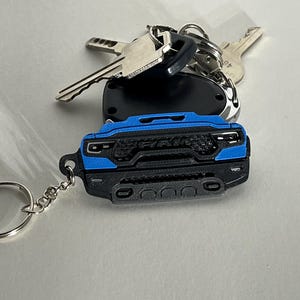 Puede incluir: Llavero negro y azul con forma de camión RAM con un llavero plateado. El llavero está sujeto a un juego de llaves y a un mando negro. El logotipo de RAM es visible en el llavero.