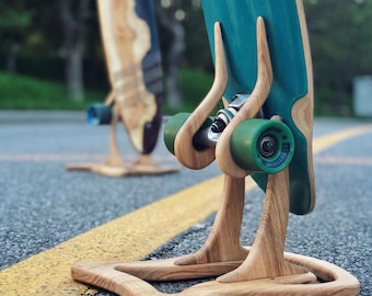 Puede incluir: Dos longboards sobre soportes de madera. Los longboards tienen cubiertas pintadas de color turquesa y azul con ruedas verdes. Los soportes son de madera clara y tienen un diseño curvo único. Las tablas se exhiben sobre una superficie pavimentada.