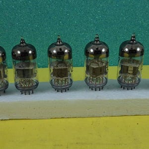 May include: Six vintage vacuum tubes with silver tops and glass bodies are arranged in a row. Each tube has internal components visible through the glass. They are set on a white rectangular base against a green and yellow background.