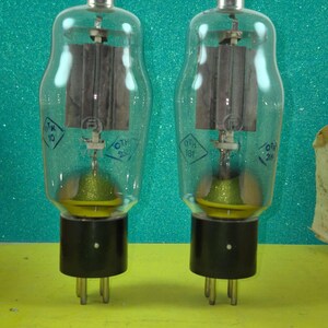 Puede incluir: Dos válvulas de vacío vintage con bombillas de vidrio transparente, componentes internos de color gris oscuro y bases negras. Los tubos tienen tapas plateadas y están etiquetados con texto y números. El fondo es un verde azulado brillante.