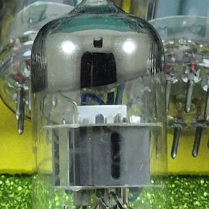 May include: A close-up of a vintage vacuum tube with a clear glass bulb and metallic components. The tube has a silver-coloured top and a series of pins at the base. The background is a glittery green.