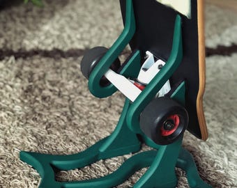 Puede incluir: Un soporte verde para monopatín con un diseño único, que sostiene un monopatín con una ilustración de dibujos animados. El soporte tiene una base decorativa y soportes curvos. El monopatín tiene ruedas negras y una tabla de madera.