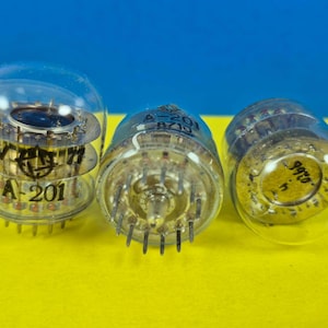 May include: Three vintage vacuum tubes with clear glass enclosures. The tubes have internal components and pins at the base. One tube displays "A-201" and other markings. The background is a bright yellow surface against a blue backdrop.