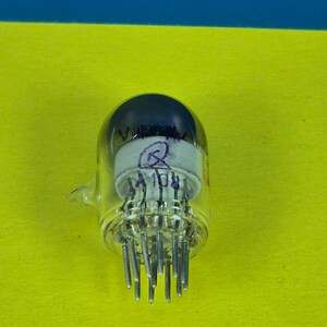 May include: A clear glass vacuum tube with a dark top and multiple metal pins extending from the base. The tube has markings in blue and red ink. The background is a bright yellow.