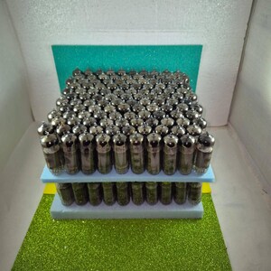 May include: A collection of vintage vacuum tubes arranged in a rectangular shape. The tubes are silver and glass, and are stacked on a blue and yellow platform, set against a green glitter background.