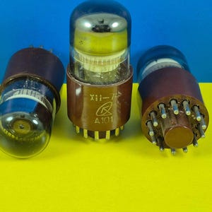 May include: Three vintage vacuum tubes with glass bulbs and brown bases are arranged on a yellow surface against a blue backdrop. The central tube has text and a logo. These electronic components are likely from older audio or radio equipment.