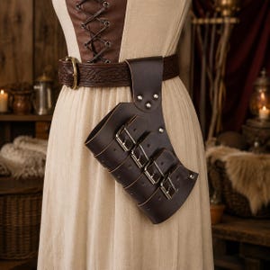 Puede incluir: Un cinturón de cuero marrón oscuro con un patrón de nudo celta en relieve y una hebilla grande de color latón. Sujeto al cinturón hay un estuche de cuero marrón oscuro estilo funda, asegurado con cuatro correas ajustables y hebillas de metal plateado.