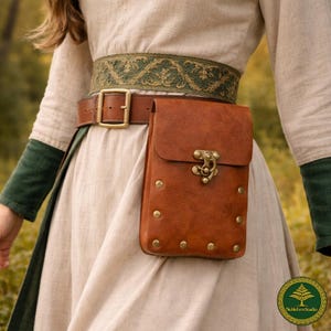 Könnte beinhalten: Eine hellbraune Ledertasche mit Messingverschluss und Metallnieten, befestigt an einem braunen Ledergürtel mit Messingschnalle. Der Gürtel wird über einem hellbeigen Leinenkleidungsstück mit einem breiten grünen Stoffbund mit Blumenmuster getragen.