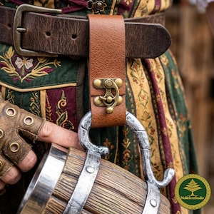 May include: A tan leather belt loop hanger featuring an antique brass latch mechanism. The hanger is attached to a dark brown leather belt and supports a wooden tankard with a silver-toned metal handle and bands, worn over a patterned garment.