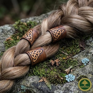 Puede incluir: Primer plano de un peinado trenzado adornado con tres cintas de cuero marrón para el cabello. Cada cinta presenta un diseño de nudo celta. La trenza descansa sobre una roca cubierta de musgo, con un fondo natural borroso.