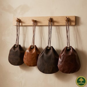 Könnte beinhalten: Vier Leder-Kordelzugbeutel in verschiedenen Brauntönen hängen an einer Holzleiste. Die Beutel haben eine abgerundete Form und sind an dünnen, braunen Kordeln aufgehängt. Die Holzleiste hat vier Holzstifte.