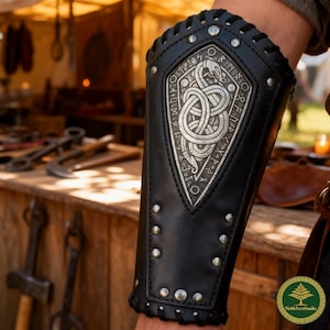 May include: A black leather forearm bracer featuring a central silver-toned metal plate embossed with an intricate serpent design surrounded by runic inscriptions. The bracer is secured with silver metal rivets and black leather lacing along the edges.