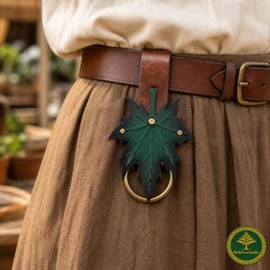 Könnte beinhalten: Ein brauner Ledergürtel mit einem hängenden Accessoire in Form eines Ahornblatts. Das Blatt ist in einem grünen und schwarzen Farbverlauf gehalten, mit einer Lederschlaufe am Gürtel befestigt und hat einen runden Messingring mit Nieten.
