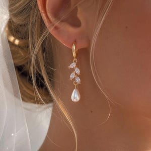 May include: A gold hoop earring featuring a vertical branch-like design adorned with three marquise-cut clear stones, finished with a single white teardrop-shaped pearl pendant hanging from the bottom of the metallic leaf-shaped arrangement.