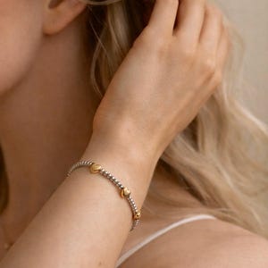 May include: A silver beaded bracelet with gold heart-shaped charms. The bracelet is worn on a wrist, with a person's arm and shoulder visible. The background is softly blurred, highlighting the jewelry.