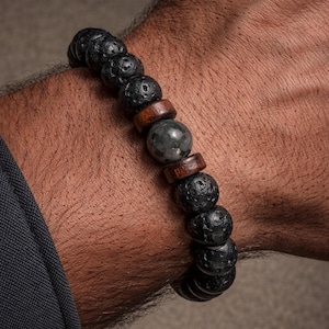 May include: A black lava stone bead bracelet with a mix of dark brown wooden beads and a single gray stone bead. The bracelet is worn on a wrist, showcasing the natural textures and colors of the materials.