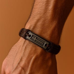 May include: A dark brown leather bracelet with a rectangular metal plate featuring a cross design. The bracelet is wrapped around the wrist, showcasing the intricate details of the religious symbol.