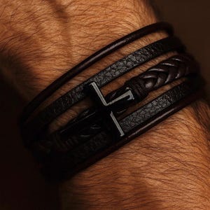 May include: Dark brown leather bracelet featuring a black cross pendant. The bracelet has multiple strands, including a braided section. The design is a mix of smooth and textured leather, creating a layered look. A stylish accessory.