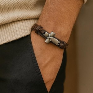 May include: A brown leather bracelet with a silver cross pendant. The bracelet is worn on a wrist, with the cross centered. The leather band is braided and secured with a clasp. The cross is a simple, classic design.