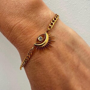 May include: Gold-toned bracelet featuring a chain link design. The bracelet has a charm with an eye-shaped design, a crescent moon, and sun rays. A small, clear gemstone is set in the eye. The bracelet is worn on a wrist.