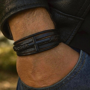 May include: Black leather bracelet with a cross design. The bracelet features multiple bands, including a braided section. The cross is a solid, dark color. The bracelet is worn on a wrist, with a black leather jacket and blue jeans in the background.