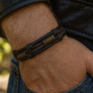 May include: A dark brown leather bracelet with bronze-colored metal accents. The bracelet features a braided design with two decorative metal cylinders. The bracelet is worn on a wrist, with a black leather jacket and blue jeans in the background.
