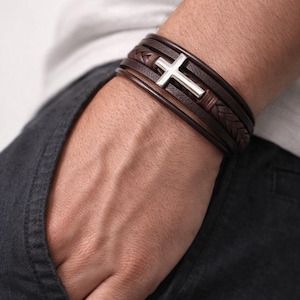 May include: A multi-layered brown leather bracelet with a silver cross charm. The bracelet features braided and smooth leather straps. The cross is centered on the bracelet, creating a simple yet elegant design.