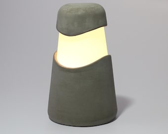 May include: A modern, gray concrete table lamp with a unique design. The lamp features a cylindrical base and a top section that partially covers the light source, creating a soft glow. The lamp's design is minimalist and contemporary.