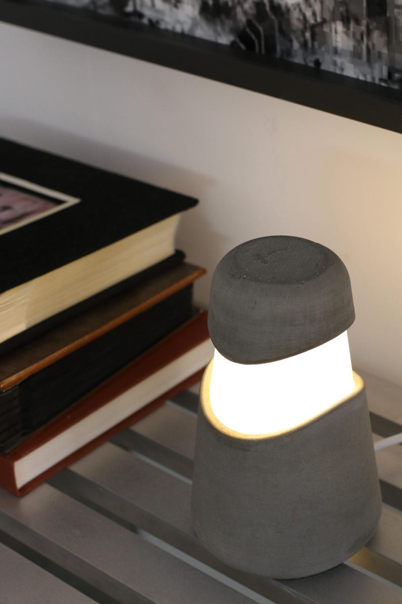 Może przedstawiać: Nowoczesna szara lampa stołowa z betonu z jasnym, świecącym wnętrzem. Lampa ma zaokrąglony wierzch i zakrzywioną, otwartą konstrukcję. W tle widoczny jest stos książek.