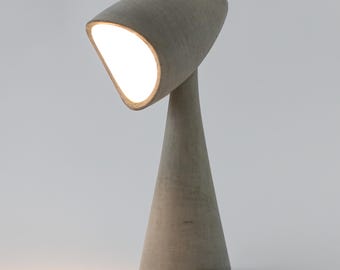 Może przedstawiać: Nowoczesna lampa biurkowa z szarego betonu, z podstawą w kształcie stożka i pochyloną, owalną głowicą świetlną. Lampa emituje jasne, ciepłe światło. Design jest minimalistyczny i rzeźbiarski, odpowiedni do nowoczesnego wnętrza. Wysokość ok. 30 cm.