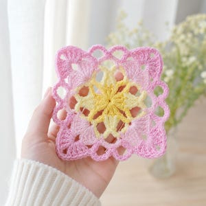 May include: A square crochet coaster featuring a central yellow star-shaped pattern surrounded by light pink and darker pink yarn. The edges of the coaster have a scalloped, open-work design with circular loops, creating a delicate, lacy appearance.