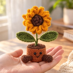 May include: A small, handcrafted crochet sunflower in a brown pot. The flower has bright yellow petals and a dark brown centre. The pot features a stitched smiling face with two black bead eyes and two small brown arms extending from the sides.
