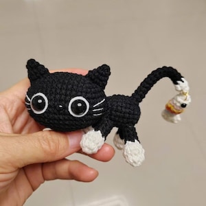 Op de afbeelding: Een handgemaakte zwart-witte gehaakte kat met grote, ronde ogen en een lange staart. De kat heeft witte poten en een kleine visbedel aan het einde van zijn staart. Het is een handgemaakt item.
