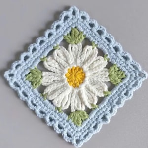 May include: A square, crocheted doily with a daisy motif. The daisy has white petals and a yellow centre, surrounded by green leaves. The border is light blue with a scalloped edge, creating a handmade, floral design.