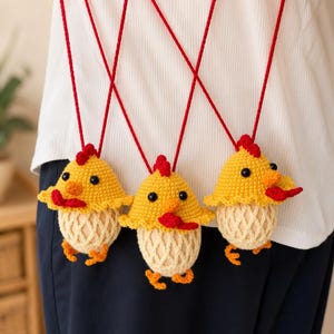 May include: Three crocheted chicken ornaments hanging from red strings. Each chicken has a yellow head with a red comb and wattle, black bead eyes, and a cream-colored body encased in a crocheted net pattern with small orange feet at the base.