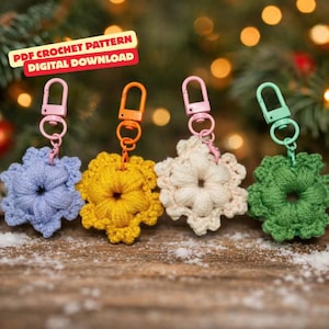 May include: Four crocheted snowflake keyrings in blue, yellow, cream, and green, each with a different coloured clasp. The keyrings are displayed on a wooden surface with a blurred background of Christmas lights and greenery. Text in the upper left corner reads "PDF Crochet Pattern Digital Download."