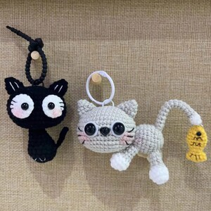 Op de afbeelding: Twee gehaakte katten sleutelhangers. E&eacute;n is zwart met grote witte ogen en roze wangen. De andere is grijs met witte poten en een gele vis. Beide hebben sleutelringen.