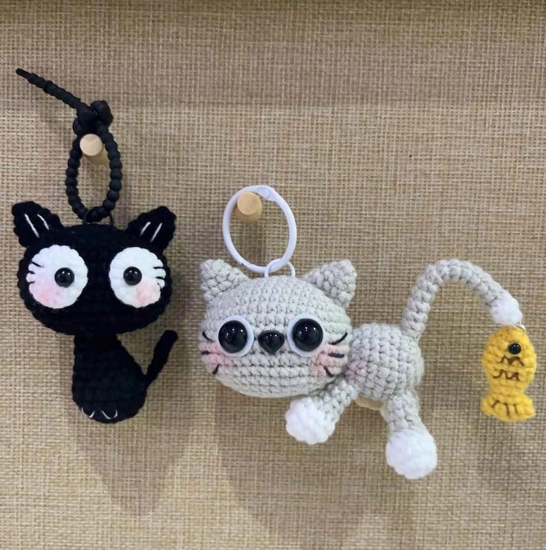 Op de afbeelding: Twee gehaakte katten sleutelhangers. E&eacute;n is zwart met grote witte ogen en roze wangen. De andere is grijs met witte poten en een gele vis. Beide hebben sleutelringen.