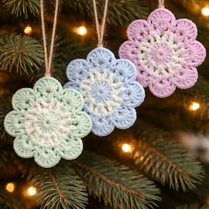May include: Three handmade crochet flower ornaments in pastel colors: green, blue, and pink. Each ornament has a white center and is suspended by a natural twine string. The ornaments are displayed on a Christmas tree.