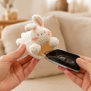 May include: A white crocheted bunny key holder with pink accents on the ears and cheeks. The bunny is holding a black car key fob. The key holder is designed to protect the key fob.