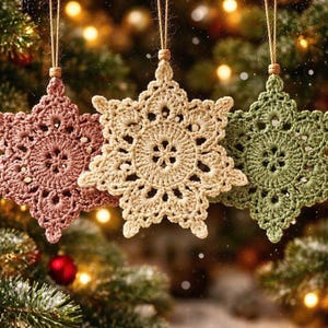 May include: Three handmade crochet snowflake decorations in pink, beige, and green hang from twine. The ornaments are in front of a blurred Christmas tree with fairy lights.