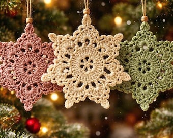 May include: Three handmade crochet snowflake ornaments in shades of pink, beige, and green hang from twine strings. The ornaments are in front of a blurred background of a Christmas tree with lights.