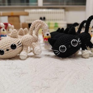 Op de afbeelding: Gehaakte kattenspeeltjes in beige en zwart. De beige kat heeft een lange, gebogen staart en de zwarte kat heeft een korte staart. Beide hebben grote, ronde ogen en witte snorharen. De speeltjes liggen op een witte, getextureerde ondergrond.