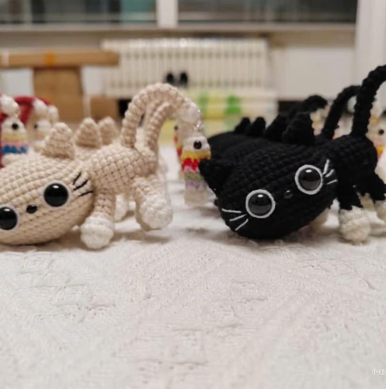 Op de afbeelding: Gehaakte kattenspeeltjes in beige en zwart. De beige kat heeft een lange, gebogen staart en de zwarte kat heeft een korte staart. Beide hebben grote, ronde ogen en witte snorharen. De speeltjes liggen op een witte, getextureerde ondergrond.