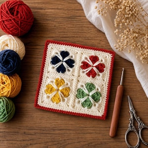 Pode incluir: Um quadrado de crochê creme com quatro desenhos florais coloridos: azul marinho, vermelho, amarelo e verde. O quadrado tem uma borda vermelha. Bolas de lã vermelha, creme, azul marinho, amarelo e verde estão ao lado, junto com uma agulha de crochê e tesouras.
