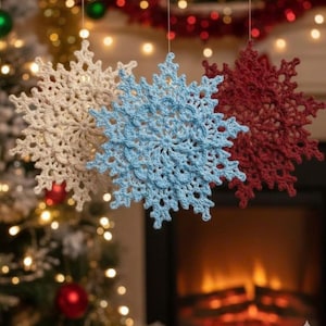 Puede incluir: Tres adornos de copos de nieve de ganchillo en crema, azul claro y burdeos cuelgan frente a una chimenea y un árbol de Navidad. Los copos de nieve tienen intrincados patrones calados, perfectos para la decoración navideña.