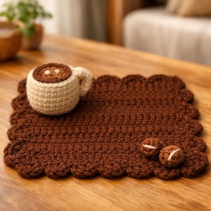 Könnte beinhalten: Ein gehäkeltes Kaffeebecher- und Untersetzer-Set. Der Becher ist beige mit braunem Innenraum, und der Untersetzer ist ein rechteckiger Rand in einem satten Braunton. Zwei kleine, runde, gehäkelte Kaffeebohnen mit weißen Akzenten sind ebenfalls vorhanden.